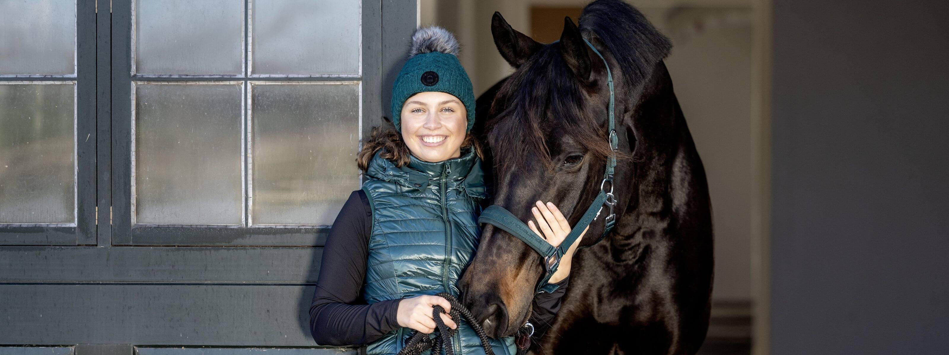 Woman in winter clothing standing next to a horse in front of a stable.