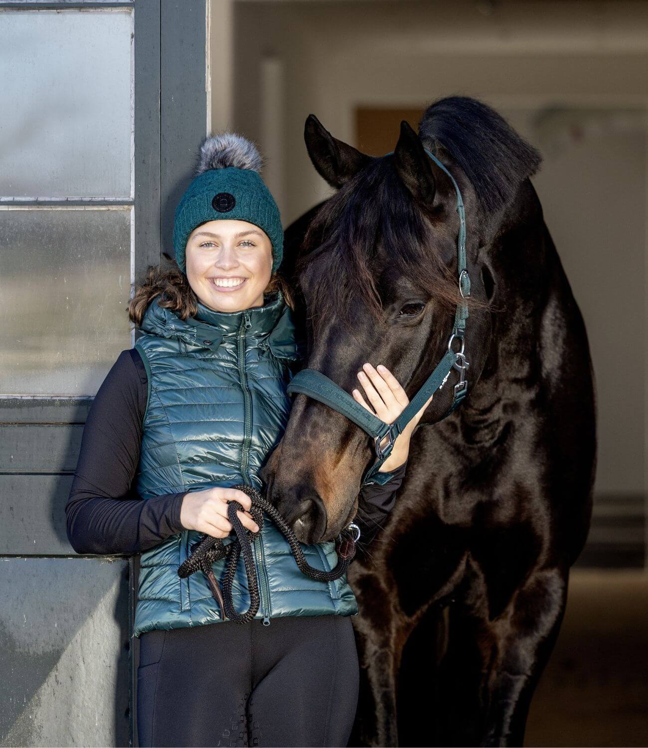 Woman in teal jacket and hat standing next to a horse, smiling.