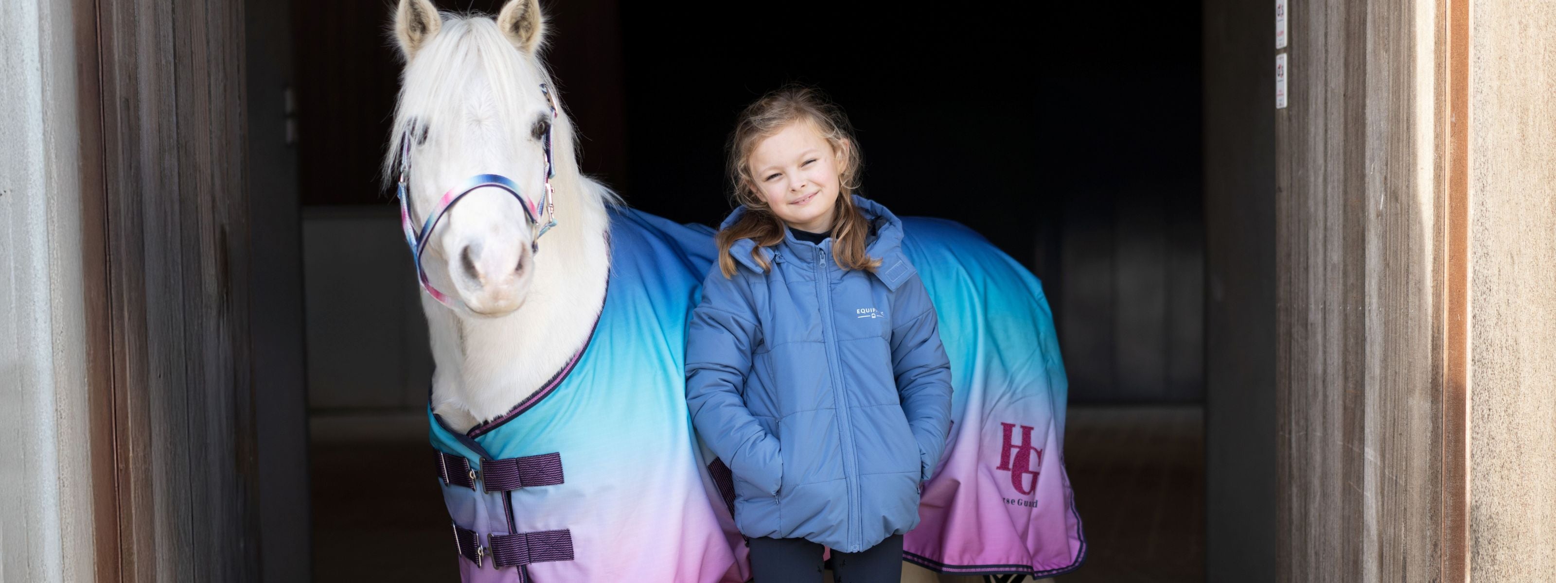 Child standing next to a horse wearing a colorful coat in a stable.