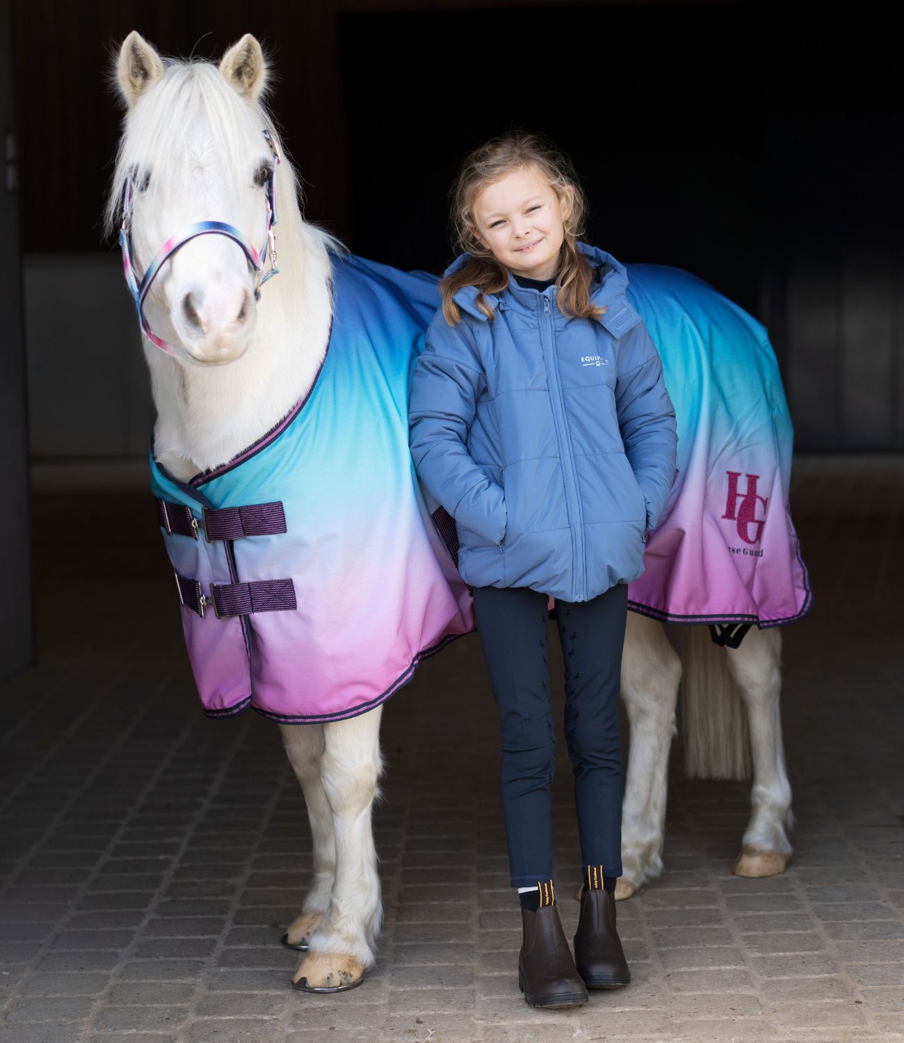Girl in equestrian attire standing next to a horse wearing a colorful rug in a stable.