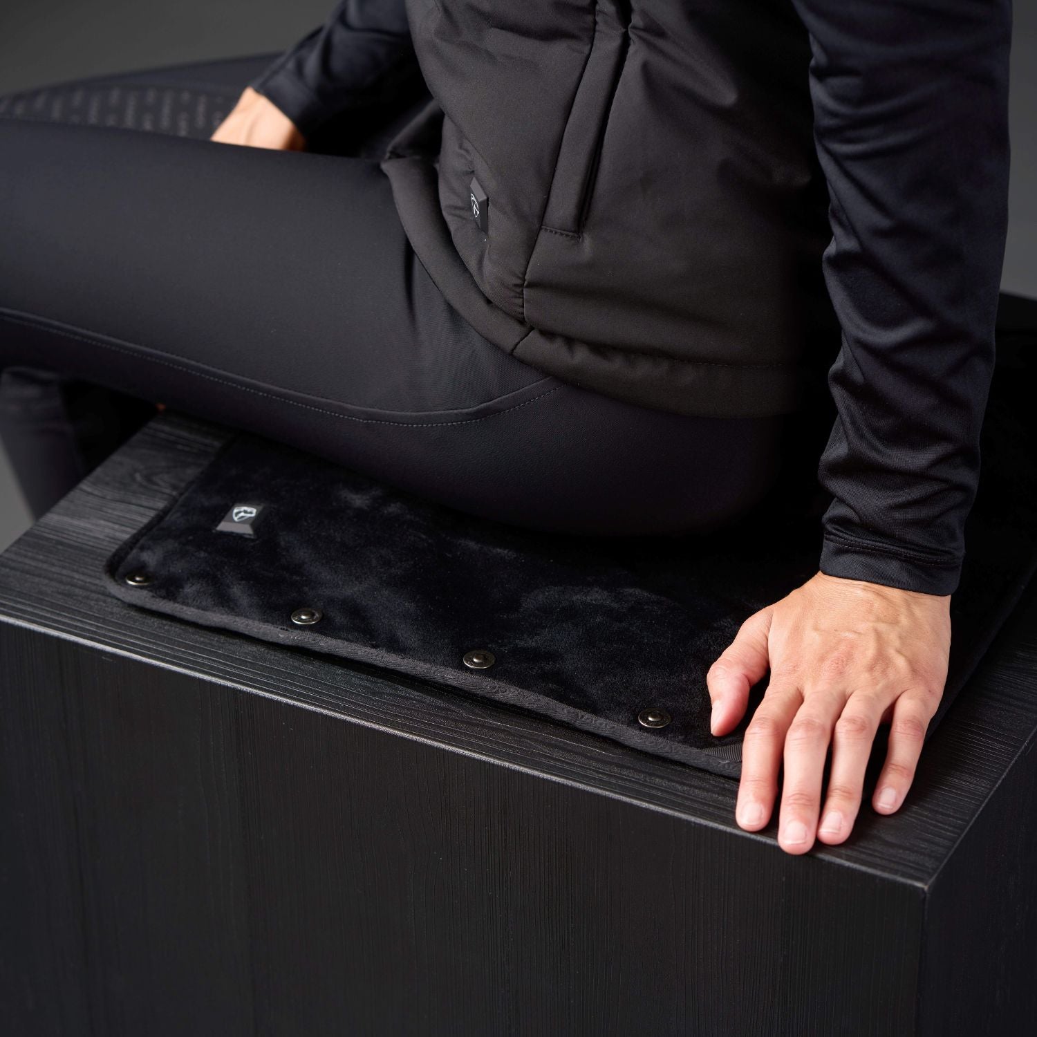 Person wearing a black outfit sitting on a black bench with a black heated mat.