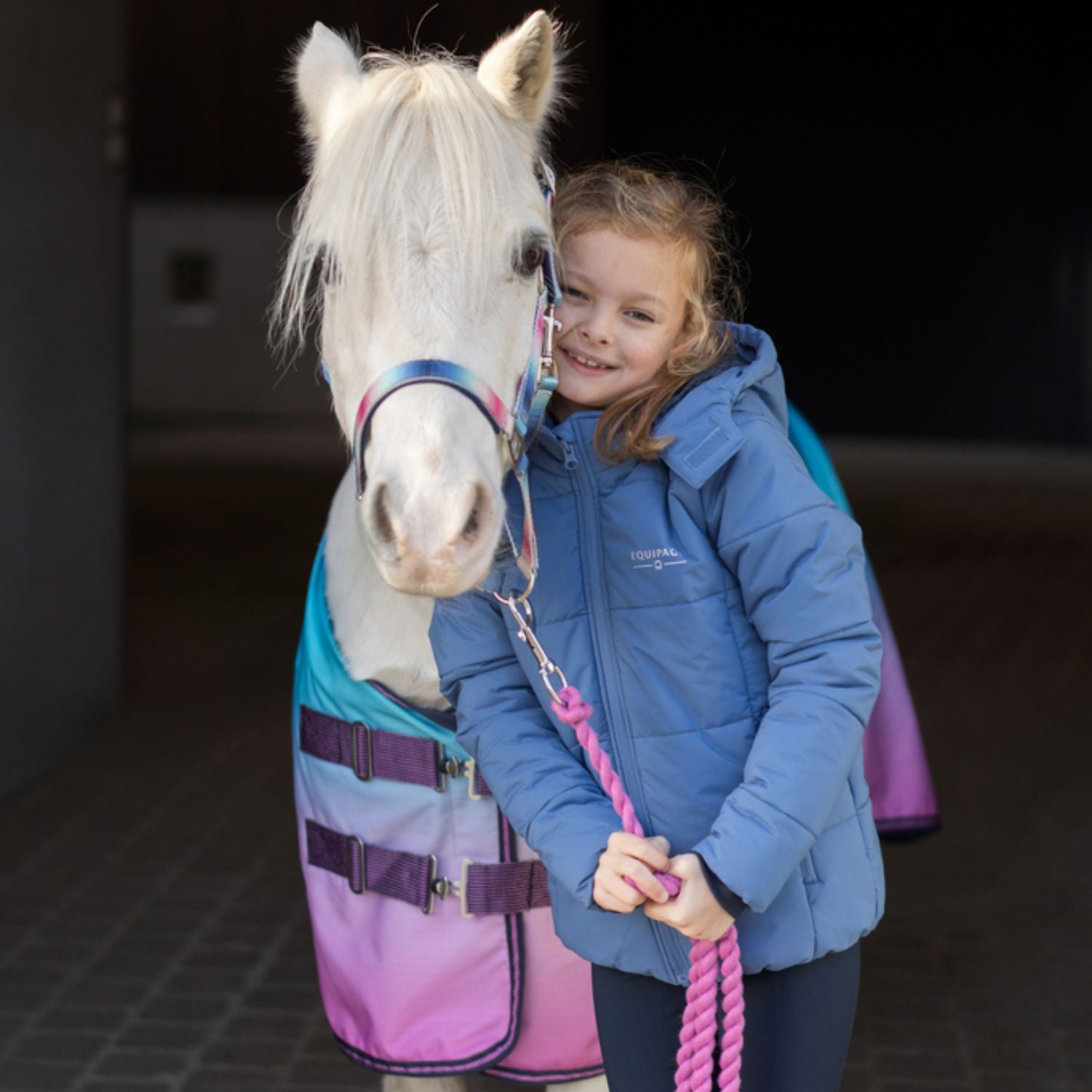 Young girl in a blue jacket holding a pink rope, standing next to a white pony wearing a colorful rug.