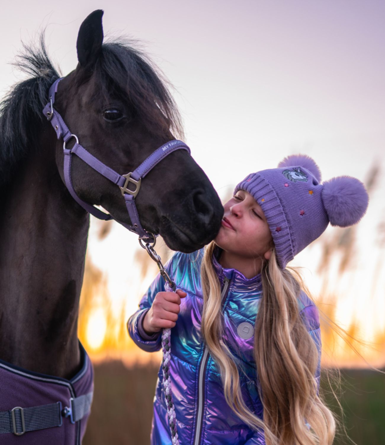 Girl in a purple outfit and hat interacting with a pony against a sunset background