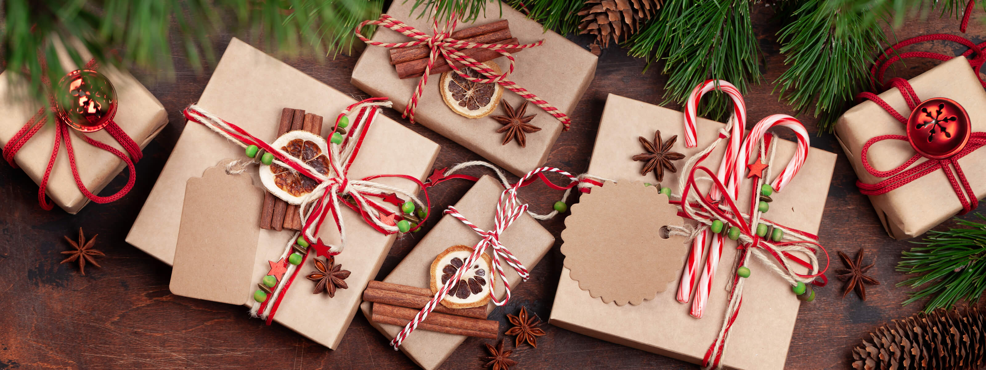 Gift boxes wrapped in brown paper with red ribbons and cinnamon sticks on a wooden surface with pine branches.