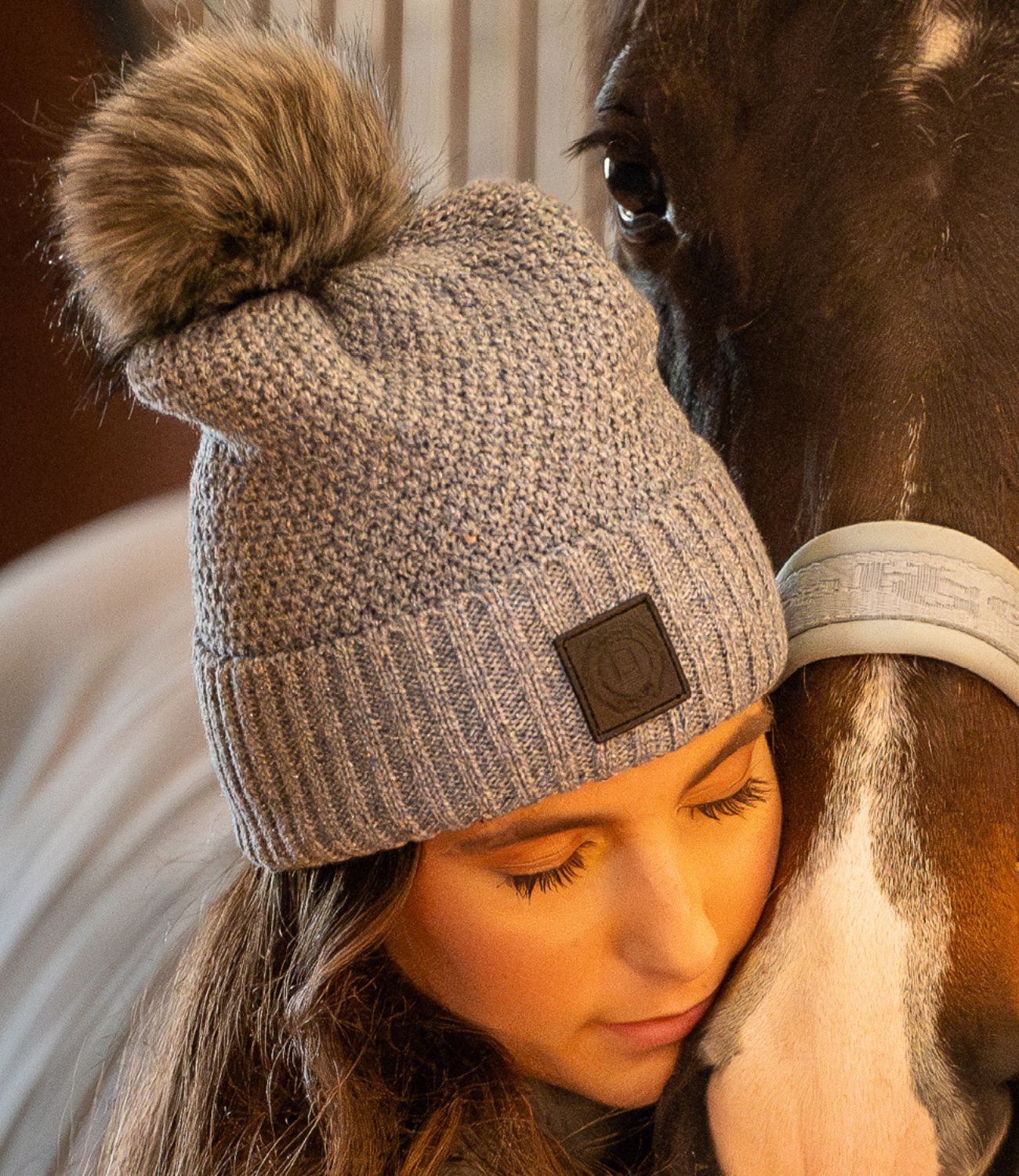 Woman wearing a gray knit beanie with a pom-pom, petting a horse.